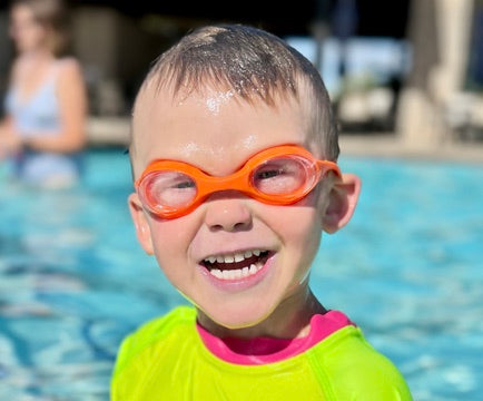 A young child wearing bright orange swim goggles and a neon yellow-green swim shirt stands in a swimming pool, smiling broadly.