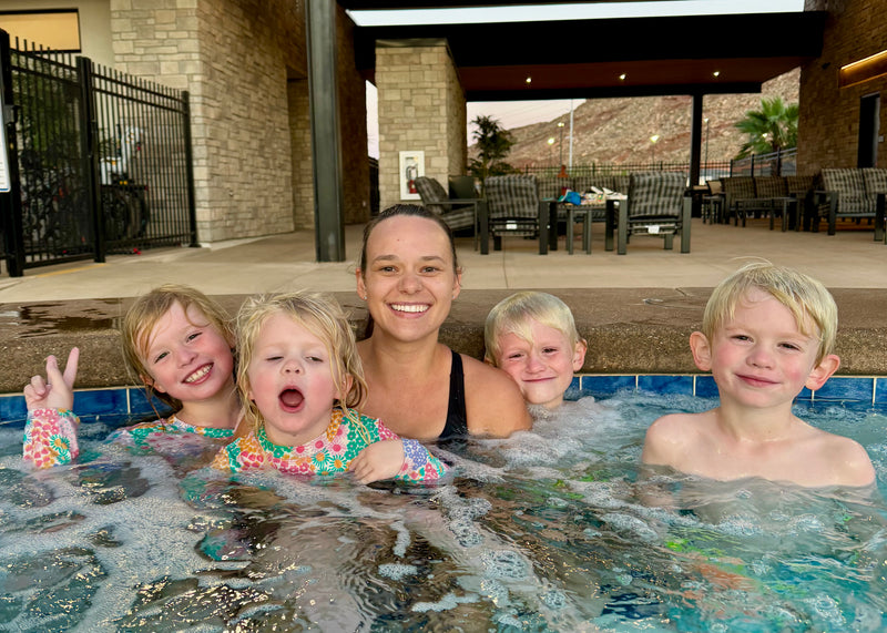 A swim teacher and four kids posing together in a bubbling outdoor hot tub pool at a resort, with a stone patio, lounge chairs, and a rocky hillside in the background.