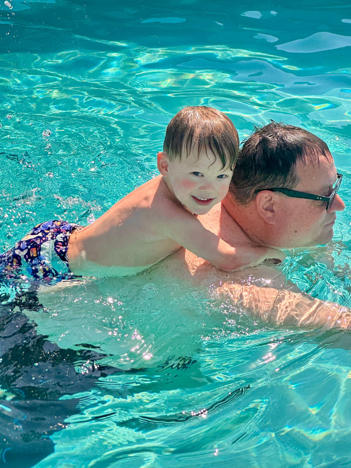 A grandpa wearing black sunglasses swims in a bright turquoise pool while holding a young child on his back. The child is wearing purple and blue patterned swim trunks and smiling at the camera.