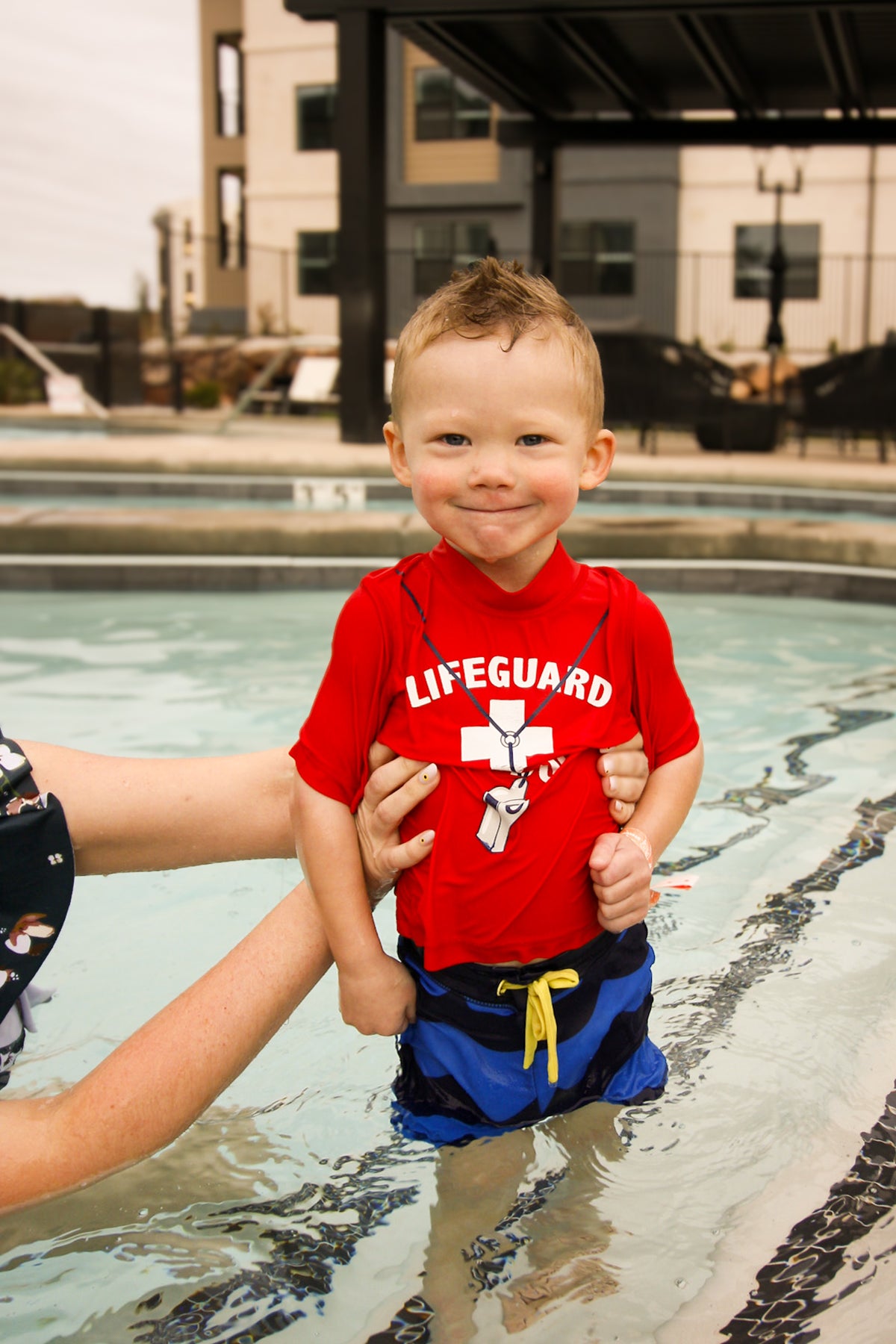 A young child standing in a swimming pool wearing a red lifeguard shirt with a white cross and yellow life vest symbol, along with blue swim trunks. An adult's arm is visible holding the child.