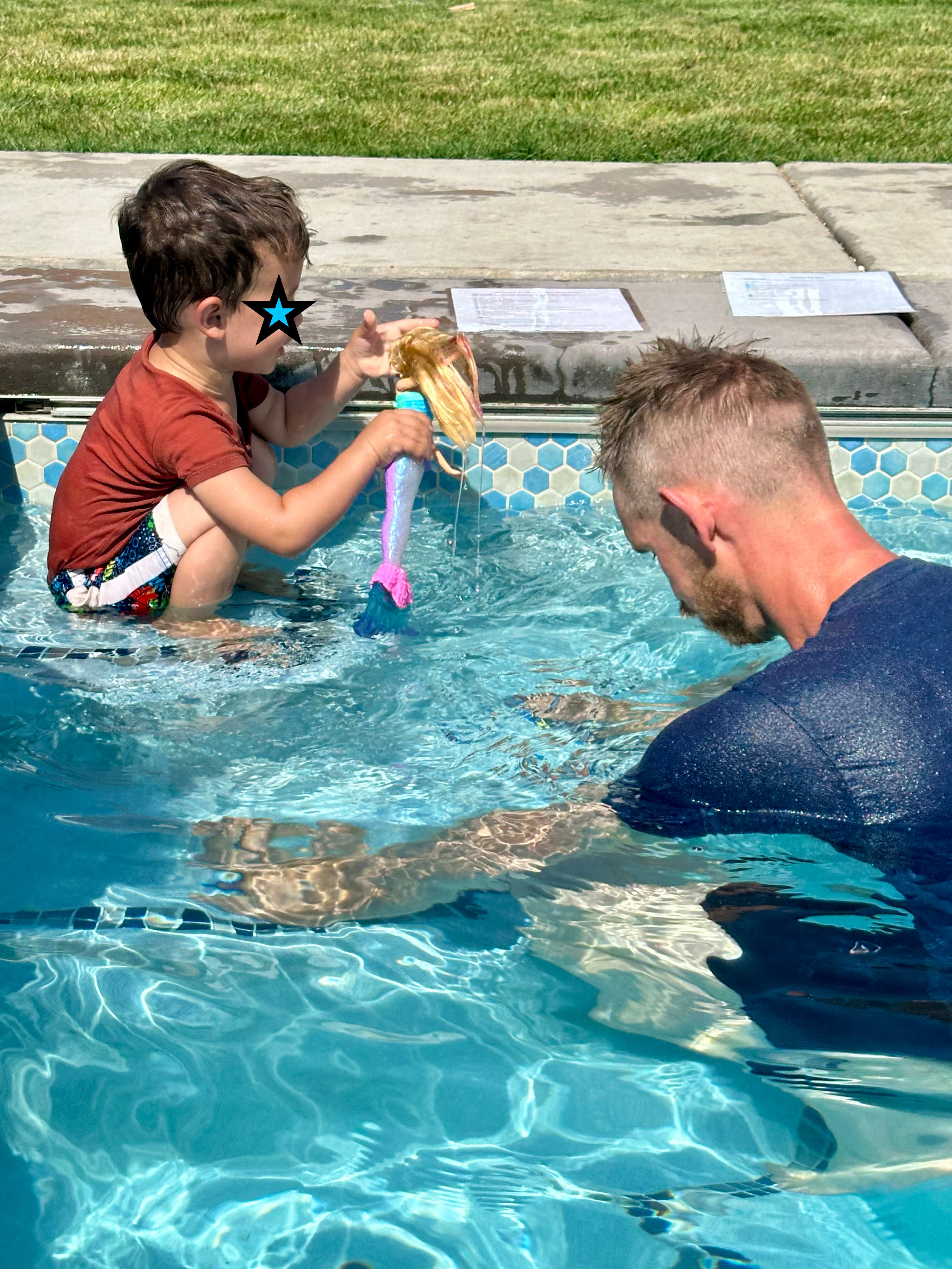 A young boy in a swimming pool wearing a red t-shirt and swim trunks, holding a toy with a long blonde wig and a pink and blue tail. An adult man in a dark blue swimsuit is sitting in the pool beside him, and a grassy lawn is visible in the background.