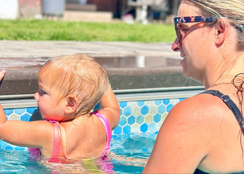 A woman in a black swimsuit and tortoiseshell sunglasses sits in a swimming pool next to a toddler wearing a pink swimsuit.