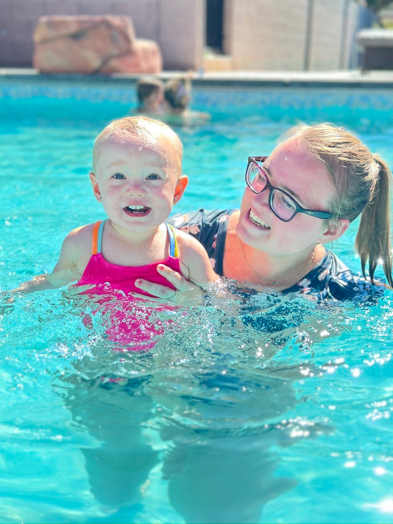 A woman wearing glasses and a navy swimsuit supports a young child in a bright pink swimsuit while swimming in a sunny outdoor pool.