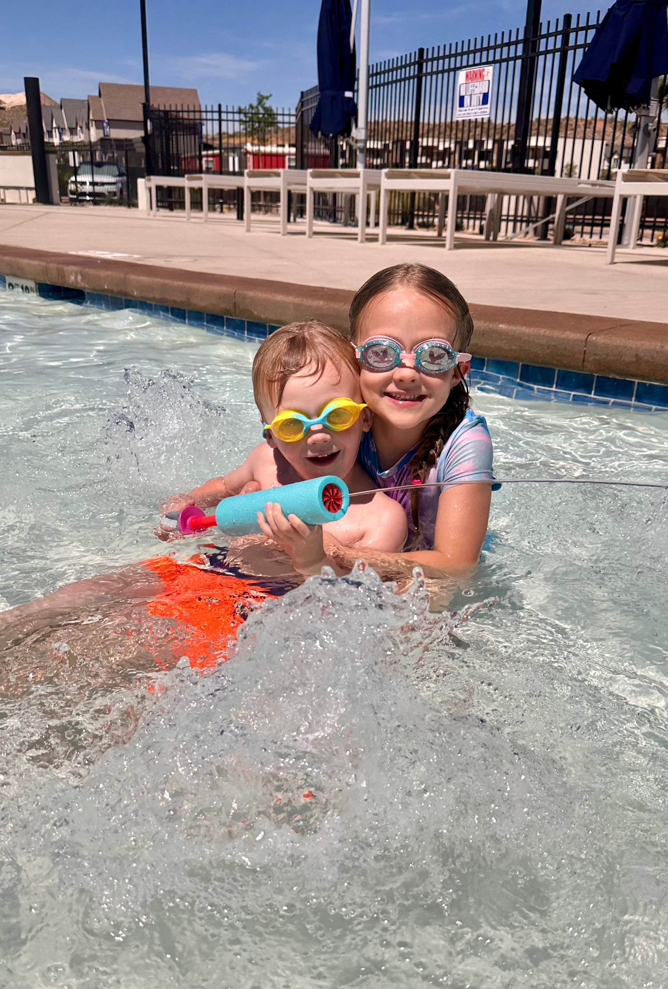 Two young children wearing swim goggles and a life vest play in a swimming pool, splashing water with a toy.